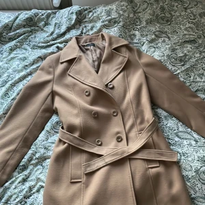 Beige kappa från Ricci Capricci - Snygg beige kappa från Ricci Capricci med klassisk dubbelknäppning och bälte i midjan. Jackan har breda slag, raka långa ärmar och två fickor framtill. Materialet är mjukt och känns lyxigt, perfekt för dig som gillar stilrena och tidlösa plagg.