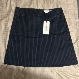 Mörkblå randig kjol Holly & Whyte - Snygg mörkblå kortkjol från Holly & Whyte med tunna vita ränder och guldfärgad dragkedja bak. Kjolen har en klassisk rak passform och är fodrad för extra komfort. 