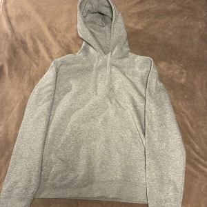 Grå hoodie med huva och snörning - En stilren grå hoodie med klassisk huva och snörning. Tröjan har en känguruficka framtill och är tillverkad i mjukt bomullsmaterial. Perfekt för chill dagar eller när du vill ha en enkel och clean look.