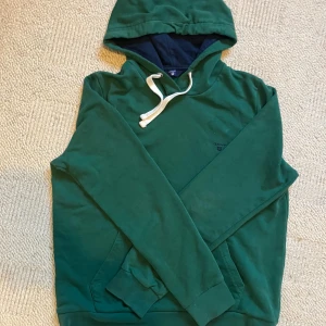 Grön hoodie från GANT med huva - Grön hoodie från GANT i storlek M med broderad logga på bröstet. Tröjan har huva med vit snörning och en klassisk känguruficka framtill. Mjuk bomullskänsla och mörkblått foder i huvan. Perfekt för chill dagar.