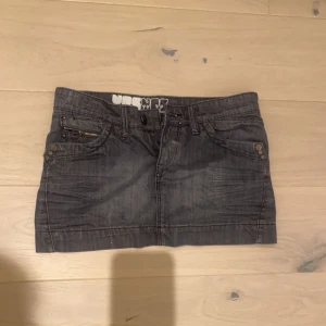 Grå jeanskjol med dragkedjor - Säljer en grå jeanskjol från UBC med kort modell. Kjolen har snygga detaljer som dragkedjor på fickorna både fram och bak, samt bälteshällor i midjan. Perfekt för dig som gillar en avslappnad och cool stil.