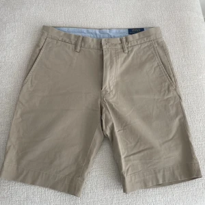 Beige shorts från Polo Ralph Lauren - Klassiska beige shorts från Polo Ralph Lauren med normal passform och midwaist. Tillverkade i mjuk bomull, har fickor i sidorna och hällor för bälte. Perfekta för sommardagar och enkel att matcha med t-shirt eller skjorta.