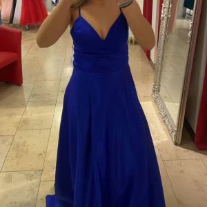 Mörkblå långklänning med snörning - Säljer en mörkblå långklänning i satin med smala axelband, v-ringad urringning och snygg snörning i ryggen. Klänningen har en elegant, figurnära överdel och ett svepande, utsvängt kjolparti. Perfekt för bal. Använd en gång ser ut som ny.