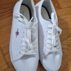 Vita sneakers från Gant i canvas - Fräscha vita sneakers från Gant i klassisk modell med lågt skaft. Skorna är i canvas med snörning och har Gant-logga broderad på sidan. Perfekta till jeans eller shorts för en clean och stilren look.