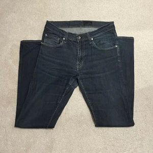 Mörkblå jeans Tiger of Sweden 32/34 - Säljer ett par mörkblå jeans från Tiger of Sweden i modellen IGGY. Klassisk femficksdesign, raka ben och snygga kontrastsömmar. Jeansen är tillverkade i ett mjukt denimtyg och har normal passform. Perfekta för dig som gillar stilrena och tidlösa jeans.