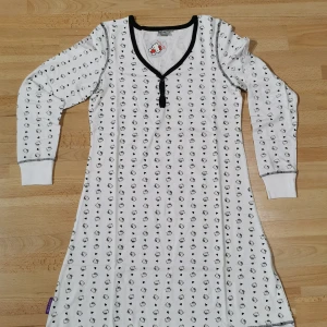 Hello Kitty vit långärmad sovplagg  - Supersöt vit långärmad sovplagg från Hello Kitty med svart Hello Kitty- och hjärtmönster över hela plagget. Sovplagg har v-ringning med svarta detaljer och knappar framtill. Tillverkad i mjuk bomull med lite stretch för extra komfort.