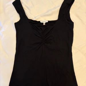 Svart v-ringad topp från Bubbleroom - Svart topp från Bubbleroom i mjuk bomull med v-ringad hals och snygg rynkning framtill. Modellen är ärmlös med breda axelband och har en slim passform som framhäver figuren. Perfekt för dig som gillar stilrena och enkla plagg med en twist.