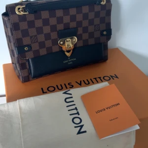 Louis Vuitton väska - Snygg brun rutig handväska från Louis Vuitton med gulddetaljer och kedjehandtag. Väskan har en elegant låsdetalj framtill och är perfekt för att ge en lyxig touch till din outfit. Köptes för 23 000kr, kvitto finns. Helt i nyskick! 