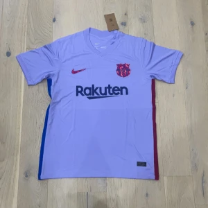 Ljuslila FC Barcelona Nike fotbollströja - Snygg FC Barcelona matchtröja från Nike i ljuslila med korta ärmar. Tröjan har klubbmärke och Nike-logga i rött på bröstet, Rakuten-tryck framtill och UNICEF-tryck på ryggen. Sidorna har blå och vinröda paneler. Tillverkad i lätt och ventilerande polyester.