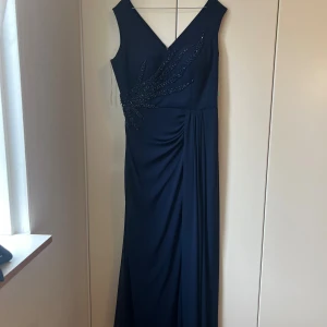 Marinblå långklänning med pärldetaljer - Elegant marinblå långklänning med v-ringning både fram och bak. Klänningen är ärmlös och har snygga pärldetaljer i ett mönster över ena sidan samt drapering vid midjan för en smickrande siluett. Perfekt för speciella tillfällen när du vill sticka ut.