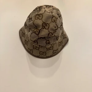 Beige bucket hat - Snygg bucket hat i beige och brunt med det klassiska monogrammet över hela hatten. Tillverkad i bomullsmix med bruna kanter och fodrad insida. Perfekt accessoar för att lyfta din outfit med lyxig känsla och streetstyle-vibe.