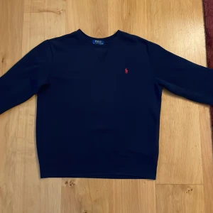 Mörkblå tröja från Polo Ralph Lauren - Säljer en stilren mörkblå tröja från Polo Ralph Lauren med klassisk röd broderad logga på bröstet. Tröjan har rund halsringning och långa ärmar, perfekt för dig som gillar enkel och tidlös design.