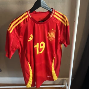 Spaniens landslagströja Lamine Yamal - Röd fotbollströja från Adidas med gula detaljer, nummer 19 och Spaniens landslagsemblem på bröstet. Namnet Lamine Yamal och nummer 19 på ryggen. Klassisk design med gula ränder på axlarna och rund hals. Tillverkad i lätt och ventilerande material.