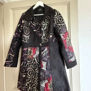 Mönstrad svart kappa från Franstyle - Unik svart kappa från Franstyle med brokigt mönster i vitt, rött och lila. Kappan har stora knappar framtill, bred krage och långärmad design. Materialet är strukturerat och känns lyxigt, perfekt för dig som gillar att sticka ut med din stil.