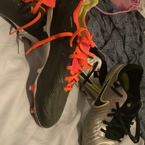 Nike Tiempo PRO & Adidas Predator fotbollsskor - Två par fotbollsskor: Nike Tiempo i silver, svart och neongul med snörning och platt sula, samt Adidas Predator i svart, vitt och rött med orange snören. Båda paren har klassisk design och är perfekta för dig som vill sticka ut på planen. Adidas stl 38 2/3 och Nike är 41. Tiempo är PRO och adidas är academy. Pris kan diskuteras. Kan gå båda för ett bättre pris😉