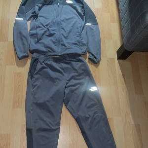 Svart vindjacka från The North Face - Säljer en svart vindjacka från The North Face med dragkedja framtill och reflekterande detaljer på ärmarna. Jackan har en sportig look och är tillverkad i lätt polyester, perfekt för träning eller chill. Klassisk logga på bröstet och normal passform. Koftan är storlek S och byxorna M