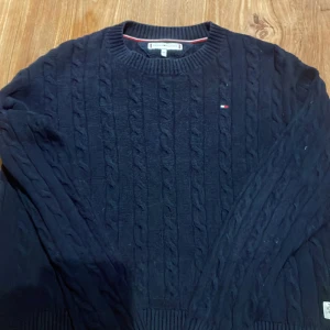 Mörkblå kabelstickad tröja Tommy Hilfiger - Kabelstickad mörkblå tröja från Tommy Hilfiger med rund halsringning och diskret logga på bröstet. Tröjan har långa ärmar och ribbade muddar vid hals, ärmslut och nederkant. Perfekt för dig som gillar klassisk och stilren look. Tappat formen efter tvätt. Pris kan diskuteras. Obs! Liten i storlek.