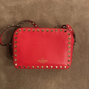 Röd axelväska från Valentino Garavani - Säljer en snygg röd axelväska från Valentino Garavani med guldiga nitar runt kanterna. Använt fåtal gånger så den är som nyskick. Säljer pga att den ej kommer till användning längre. Skriv privat för fler frågor 