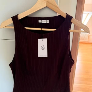 Mörkröd ärmlös klänning Stradivarius - Snygg ärmlös klänning från Stradivarius i mörkröd nyans med tunna vertikala ränder. Modellen är figurnära med rund halsringning och diskret dragkedja i sidan. Perfekt för dig som gillar stilrena och trendiga plagg.