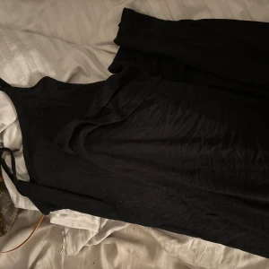 Svart långklänning från Gina Tricot - En svart långklänning från Gina Tricot i enkel och stilren design. Klänningen har smala axelband och är figurnära med rak passform. Perfekt för dig som gillar minimalistisk stil och vill ha en klassisk svart klänning i garderoben.