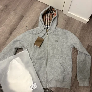 Grå hoodie från Burberry med dragkedja - Grå hoodie från Burberry med klassiskt rutigt foder i huvan, dragkedja framtill och snörning vid huvan. Tröjan har två fickor och broderad logga på bröstet. Perfekt för en chill och stilren look.