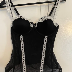 Svart korsett topp  - Säljer en svart korsett topp i storleken S (men hur tajt den sitter går att justera) med vita spetsdetaljer och tunna axelband. Materialet är tunt och ger en snygg siluett. Perfekt party topp.
