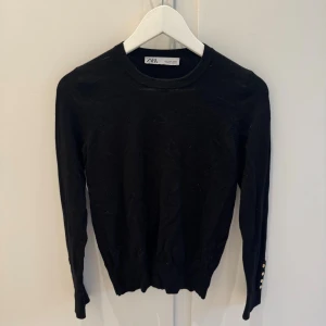Svart långärmad tröja från Zara - Svart långärmad tröja från Zara med rund halsringning och snygga guldknappar vid ärmsluten. Tröjan är i mjukt material och har en klassisk, stilren look som passar perfekt till jeans eller kjol.