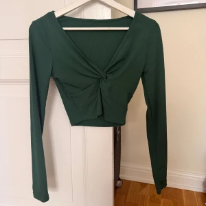 Grön croppad topp med twist  - Mörkgrön croppad topp med lång ärm och snygg twistdetalj framtill. Toppen är ribbad och har v-ringning, vilket ger en trendig och cool look. Perfekt att styla med höga jeans eller kjol.