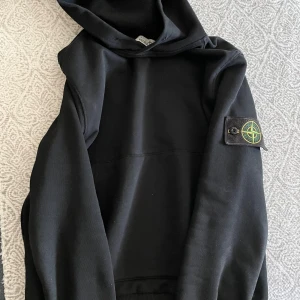 Svart hoodie från Stone Island - Svart hoodie från Stone Island med klassisk patch på vänster ärm. Tröjan har huva och långa ärmar, tillverkad i mjuk bomull. Perfekt för dig som gillar streetwear och vill ha en stilren look med ikonisk detalj.