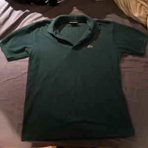 Mörkgrön pikétröja från Lacoste XL - Säljer en klassisk mörkgrön pikétröja från Lacoste i storlek XL. Tröjan har korta ärmar, krage och knäppning framtill. Ikoniska krokodilloggan är broderad på bröstet. Perfekt för dig som gillar stilren och sportig look.