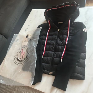 Moncler cardigan svart - Svart pufferjacka från Moncler med stickade ärmar och quiltad front. Jackan har huva med röd, vit och blå snörning, dragkedja framtill och Moncler-logga på ärmen. Materialmix av polyester och stickad textil, perfekt för dig som vill ha en snygg streetstil. Skulle säga kan vara kort i kroppen fast ganska bra längd i armarna.