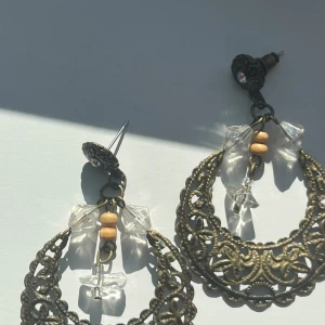 Vintageinspirerade örhängen med pärlor - Unika örhängen i antikguld med utskurna detaljer, dekorerade med klara plastpärlor och små träpärlor. Halvmåneformad design med boho-vibe, perfekt för dig som gillar statement-smycken. Fäste med liten glittrig sten för extra detalj.