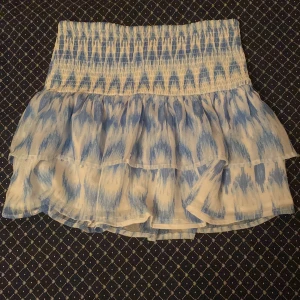 Blåvit volangkjol från Gina Tricot - Supersöt kort kjol från Gina Tricot i blått och vitt med volanger och smock i midjan. Kjolen har ett fint blått mönster, den är tunn och perfekt för varma dagar. Passar dig som gillar en feminin och trendig stil.