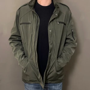 Olivgrön vindjacka med dragkedjor - Snygg olivgrön vindjacka med flera praktiska fickor och dragkedjor på bröstet och ärmen. Jackan har hög krage, knapp- och dragkedjestängning samt en clean, sportig look. Perfekt för dig som gillar stilren och funktionell design.