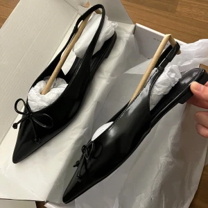 Svarta ballerinaskor med spetsig tå - Snygga svarta ballerinaskor i skinn med spetsig tå och öppen häl. Skorna har en elegant rosettdetalj framtill och platt sula, perfekta för en stilren look. Passar till både jeans och klänning. Helt nya i storleken 36, från NAKD.