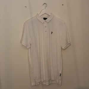 Vit pikétröja med liten logga - Klassisk vit pikétröja från Peak Performance med korta ärmar och krage. Tröjan har tre knappar framtill och en liten svart logga broderad på bröstet. Perfekt för en clean och sportig look.