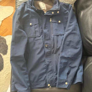 Blå vindjacka från J.Lindeberg - Snygg marinblå jacka från J.Lindeberg med hög krage, flera fickor med tryckknappar och dragkedja framtill. Jackan har ett diskret logomärke på knapparna och är tillverkad i lätt polyester. Perfekt för dig som gillar stilren och sportig look. Priset går definitivt att diskutera