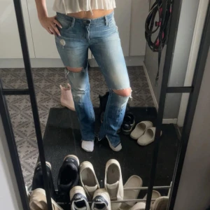 Ljusblå slitna bootcut jeans - Säljer ett par ljusblå jeans med slitningar och hål vid knäna. Modellen har bootcut-ben och klassisk femficksdesign. Jeansen har låg midja och är tillverkade i denim med snygg tvätt och slitna detaljer för en cool look.