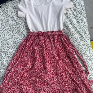 Röd blommig klänning med knytband - Säljer en röd långkjol med vitt blommigt mönster och knytband i midjan. Kjolen har en luftig och flowy passform, perfekt för dig som gillar färg och mönster. Materialet känns mjukt och lätt, troligen bomull eller polyester. Supersnygg till en enkel topp!