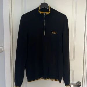 Svart Hugo Boss half zip tröja - Svart långärmad tröja från Hugo Boss med gul logga på bröstet och gul detalj vid krage och muddar. Half zip framtill och ribbad krage. Perfekt för dig som gillar stilrena och sportiga plagg med snygga kontrastdetaljer.