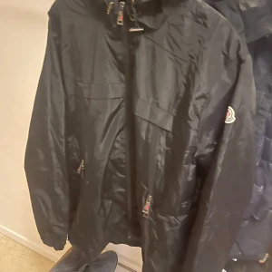 Svart Moncler vindjacka - Svart vindjacka från Moncler i storlek XXL. Jackan har dragkedja framtill, två fickor med dragkedja och huva med Moncler-logga broderad längs kanten. Tillverkad i glansig polyester och har en cool serietidningsdetalj på insidan.