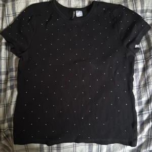 Svart ribbad topp med pärlor Divided - Svart ribbad topp från Divided med korta ärmar och små pärlor över hela framsidan. Toppen har en klassisk rund halsringning och är gjord i ett stretchigt material som sitter snyggt på kroppen. Perfekt för att lyfta en enkel outfit.