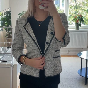 Grå kavaj med svarta detaljer - Säljer en snygg gråmelerad kavaj med svarta kanter och dekorativa knappar. Jackan har trekvartsärm och två fickor framtill med svarta detaljer.