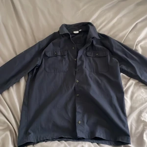 Mörkblå overshirt från Name It - Snygg mörkblå skjorta från Name It med klassisk krage och två bröstfickor med knappar. Skjortan har lång ärm och knäppning hela vägen framtill. Perfekt för dig som gillar enkel och stilren design. Passar dig som är 13-14 år.