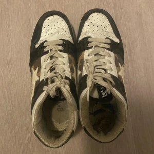 Bape Sta sneakers med camomönster - (inuti är det lite trasig men man kan ersätta fot sulan till någon annat!!) Säljer ett par Bape Sta sneakers i vitt, svart och beige med ikoniskt camomönster på sidorna och den klassiska stjärnan. Skorna har snörning, rund tå och platt sula. Perfekta för dig som gillar streetwear och vill sticka ut med en unik look.
