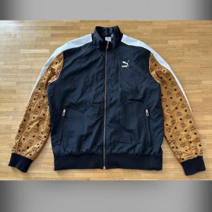 Puma x MCM vindjacka - Snygg vindjacka från Puma i samarbete med MCM. Jackan är svart med bruna ärmar i MCM:s klassiska monogram och vita ränder längs axlarna. Dragkedja framtill, ståkrage och broderad MCM-logga på ryggen. Perfekt för dig som gillar streetwear och unika plagg. 