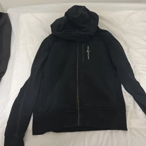 Svart hoodie från Sail Racing - Svart hoodie från Sail Racing med dragkedja framtill och en extra dragkedjeficka på bröstet. Broderad logga på vänster sida. Tröjan har huva och är tillverkad i mjukt material, perfekt för kyliga dagar.