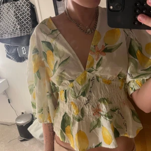 Blommig blus med citronmönster - Superfin blus med puffiga långärmar och ett färgglatt citron- och blommönster på vit botten. Toppen har smockad midja som ger en snygg form och voluminösa ärmar. Perfekt för dig som vill sticka ut med en fräsch och somrig vibe. Storlek m men passar min som vanligtvis har s/xs. Helt ny med prislapp kvar endast provad💓