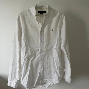 Vit skjorta från Ralph Lauren - Snygg vit skjorta från Ralph Lauren i slim fit med klassisk krage och knappar framtill. Stretchig bomull som sitter skönt och har broderad logga på bröstet. Perfekt för dig som gillar stilrena plagg. Modellen är 180cm