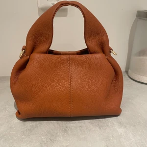 Brun handväska - Snygg och trendig handväska i fin brun färg, med guldfärgade metalldetaljer. Väskan är helt oanvänd och original priset är 1499kr. Med väskan kommer även justerbar axelrem. 